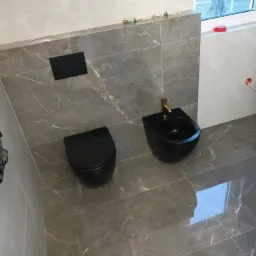 Wykończona łazienka z szarymi płytkami imitującymi marmur, wisząca czarna toaleta i bidet z złotą armaturą, widoczny grzejnik drabinkowy i okno.