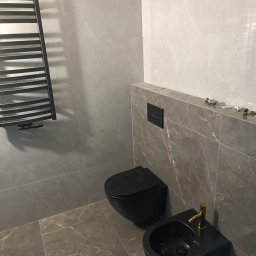 PAWEŁ MOŚCICKI - Nowoczesna łazienka w szarościach z czarną armaturą: wisząca toaleta, bidet z złotą baterią oraz grzejnik drabinkowy na tle marmurowych płytek. Widoczne niezakończone prace instalacyjne.