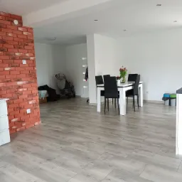 Jasne panele podłogowe ułożone w przestronnym wnętrzu z ceglanym narożnikiem, białe ściany i stół z krzesłami w tle.