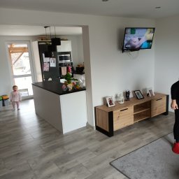 PAWEŁ MOŚCICKI - Jasne wnętrze domu z szarymi panelami podłogowymi, wyspą kuchenną z czarnym blatem, szafką RTV z dekoracjami i telewizorem na ścianie, oraz małą dziewczynką w tle przy drzwiach balkonowych.