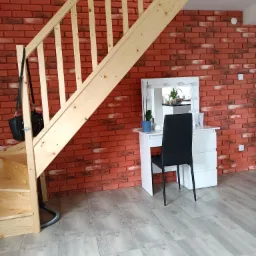 Drewniane schody z czarną torbą na poręczy, czerwona ściana z cegły i biała toaletka z lustrem oświetlonym lampkami, szare panele podłogowe.