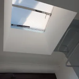 Widok z dołu na otwarte okno dachowe z żaluzjami w białej obudowie, widoczny fragment grzejnika i okna w ścianie bocznej.