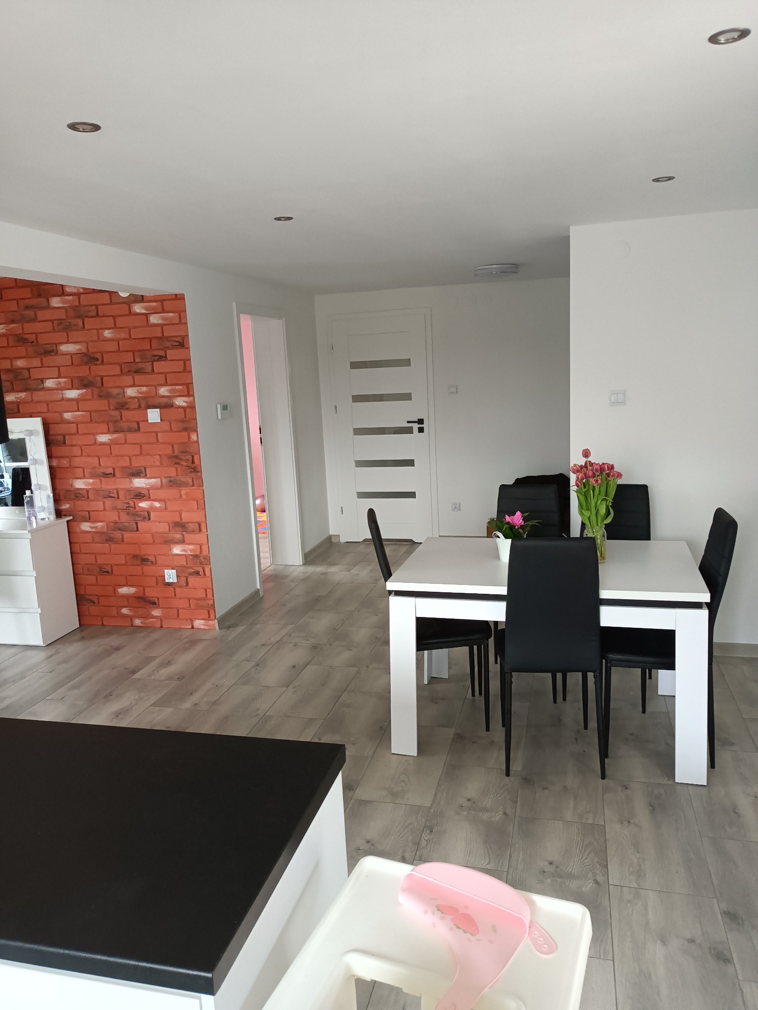 Jasne wnętrze jadalni z białym stołem, czarnymi krzesłami, bukietem tulipanów i podłogą imitującą drewno, z fragmentem ściany wykończonej czerwoną cegłą dekoracyjną