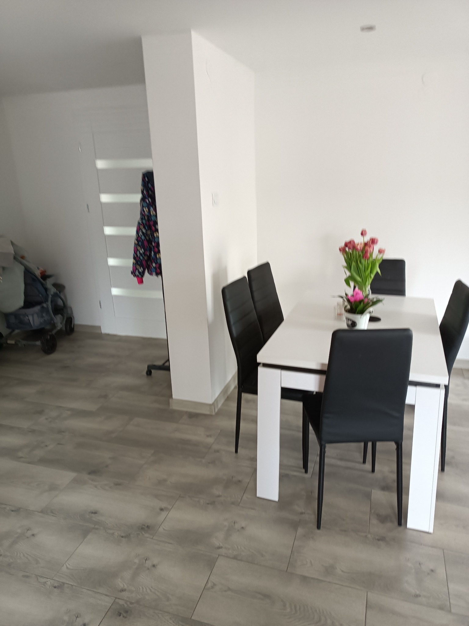 Jasne wnętrze jadalni z białym stołem, czarnymi krzesłami i bukietem tulipanów na blacie, podłoga wyłożona panelami imitującymi drewno.