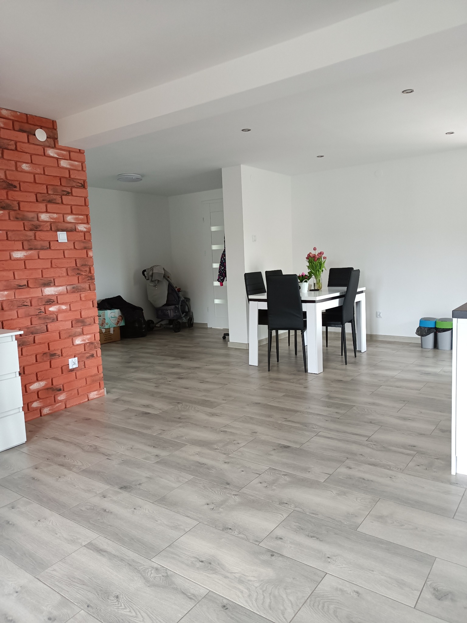 Jasne panele podłogowe ułożone w przestronnym wnętrzu z ceglanym narożnikiem, białe ściany i stół z krzesłami w tle.