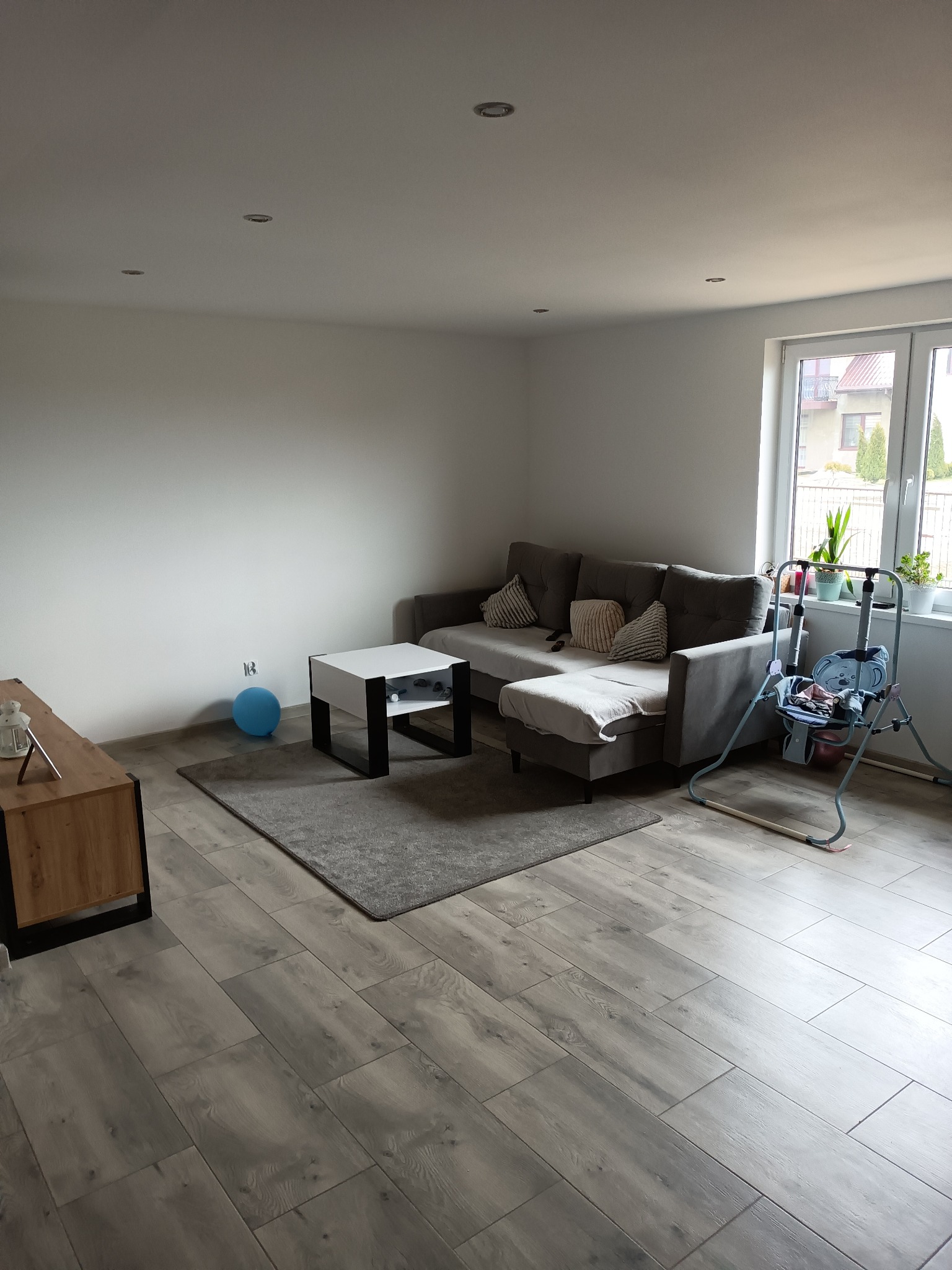 Jasne, przestronne wnętrze salonu z szarą sofą narożną, stolikiem kawowym, drewnianą podłogą, niebieskim balonem przy ścianie i huśtawką dla dziecka przy oknie.