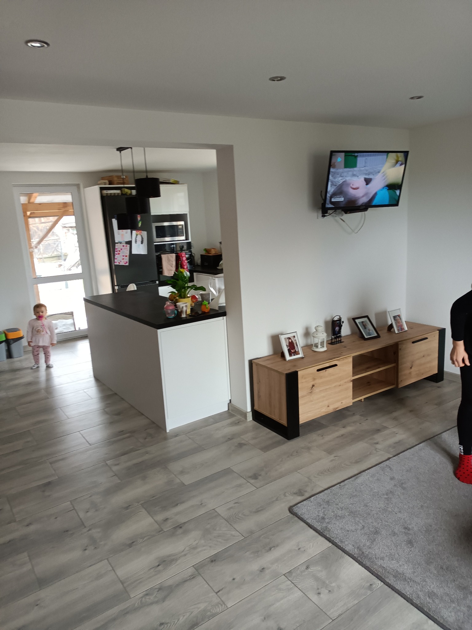 Jasne wnętrze domu z szarymi panelami podłogowymi, wyspą kuchenną z czarnym blatem, szafką RTV z dekoracjami i telewizorem na ścianie, oraz małą dziewczynką w tle przy drzwiach balkonowych.