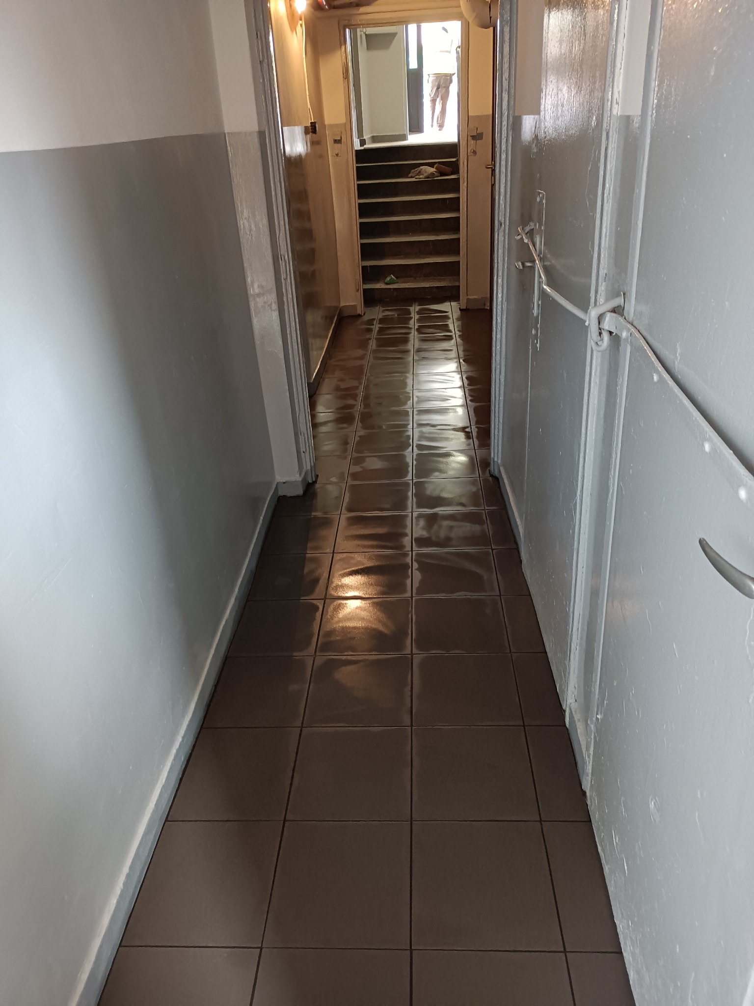 Korytarz w bloku z szaro-białymi ścianami, brązowymi błyszczącymi płytkami na podłodze i widocznymi schodami w tle.