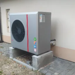 Szara pompa ciepła powietrze-woda z czerwoną linią, zainstalowana na betonowych bloczkach przy ścianie budynku z kostką brukową i opadłymi liśćmi.