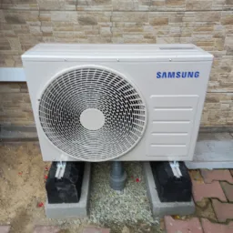 Zewnętrzna jednostka klimatyzacji Samsung zamontowana na betonowych bloczkach i gumowych podkładkach, z widoczną rurą odprowadzającą skropliny, na tle muru z cegły.