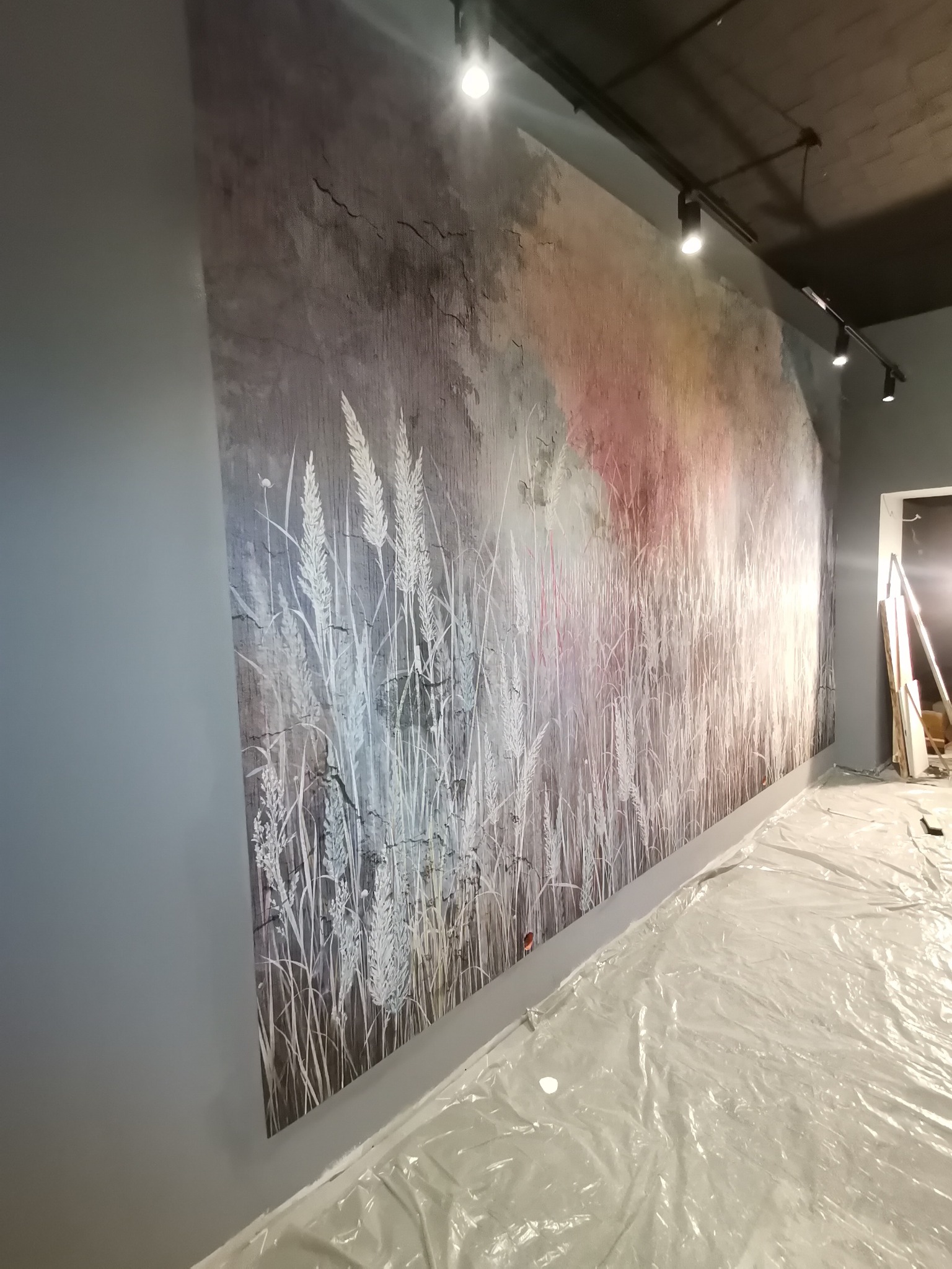Ściana z szarą tapetą w motywie łąki z białymi źdźbłami, podłoga zabezpieczona folią malarską, oświetlenie punktowe sufitowe.