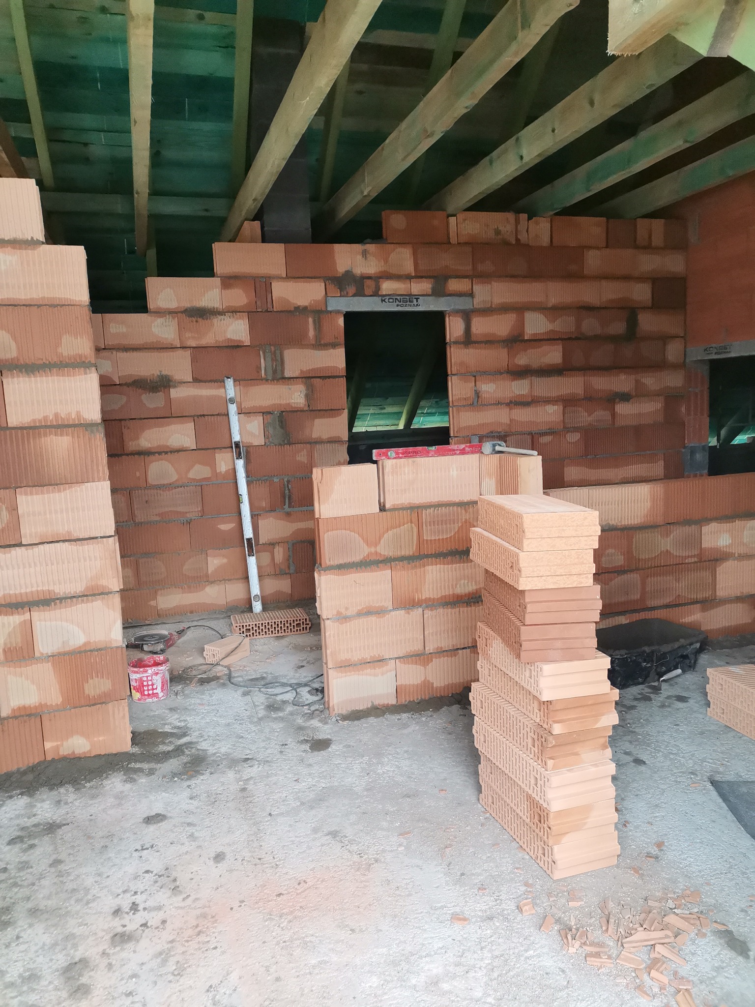 Wznoszenie muru z czerwonej cegły ceramicznej, widoczne drewniane belki stropowe, poziomica oparta o ścianę, narzędzia i zapas cegieł na podłodze w surowym stanie.