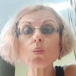 Portret kobiety z blond włosami i okularami, robiącej dzióbek ustami, z widocznymi zmarszczkami na czole i delikatnym makijażem oczu.