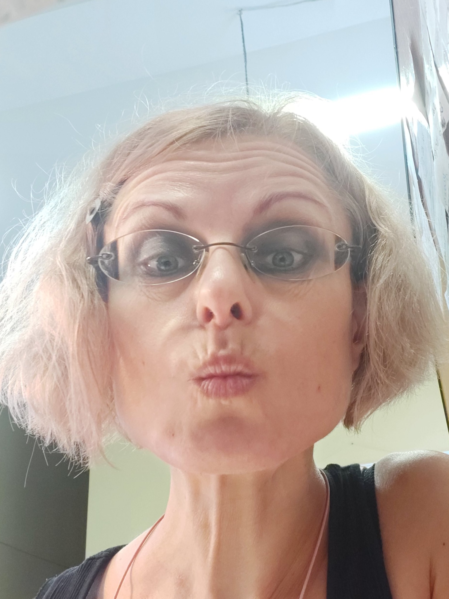 Portret kobiety z blond włosami i okularami, robiącej dzióbek ustami, z widocznymi zmarszczkami na czole i delikatnym makijażem oczu.