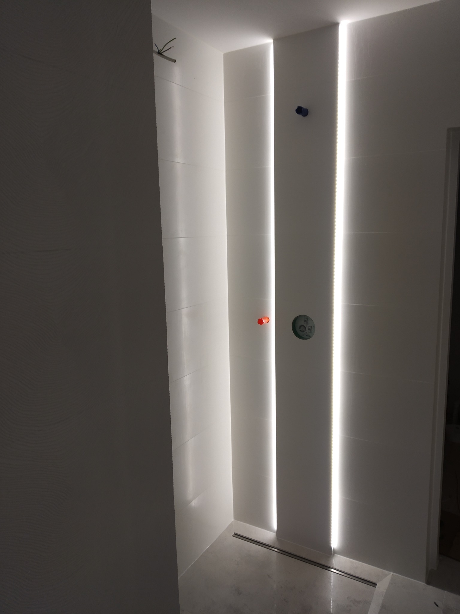 Nowoczesna łazienka z białymi płytkami, oświetleniem LED w pionowych pasach i minimalistycznym odpływem liniowym. Widok na fragment ściany i podłogi.