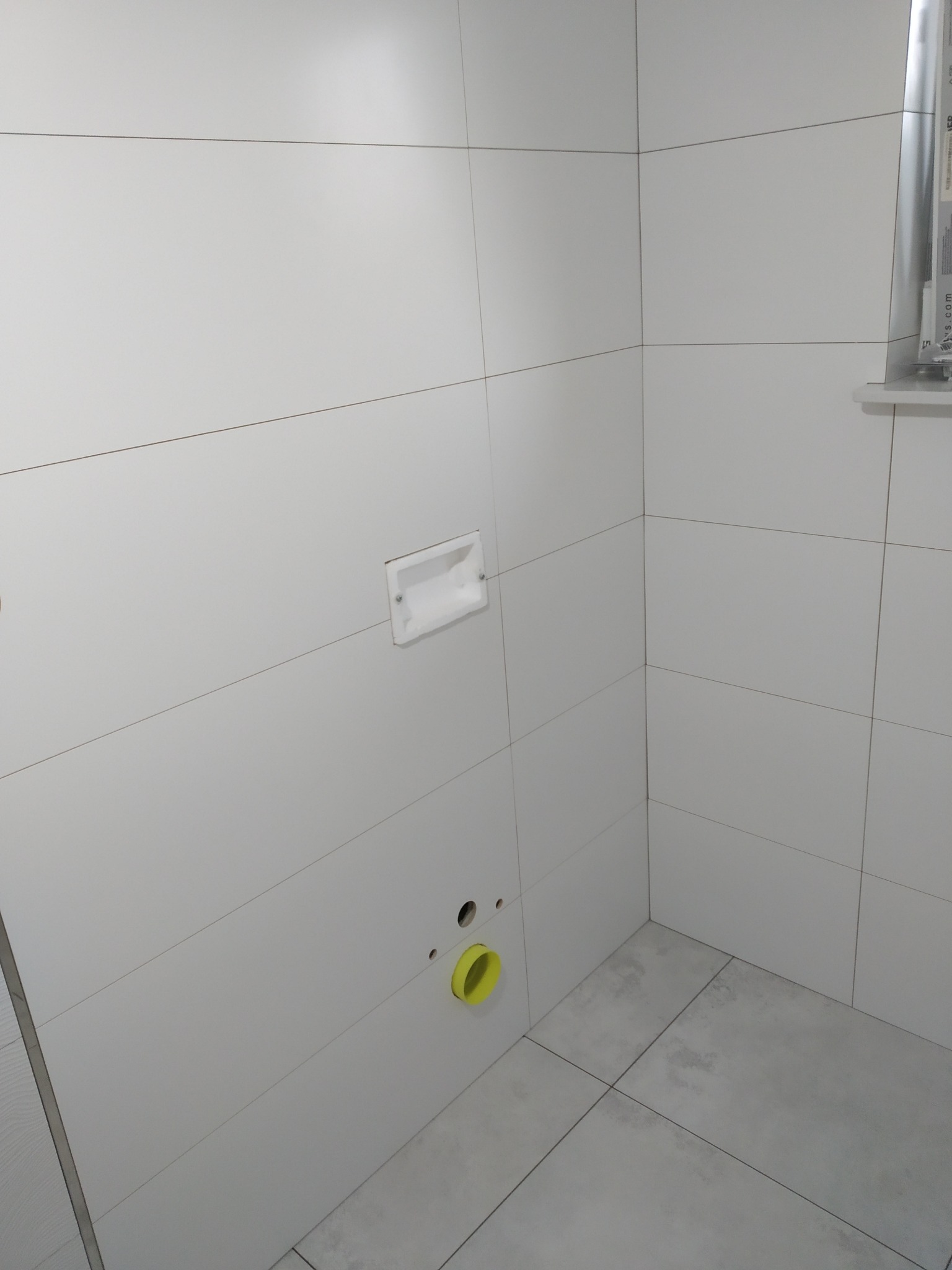 Wykończona łazienka z białymi, poziomymi płytkami na ścianach i szarymi na podłodze. Widoczne otwory na rury i uchwyt na papier toaletowy. Minimalistyczny design.