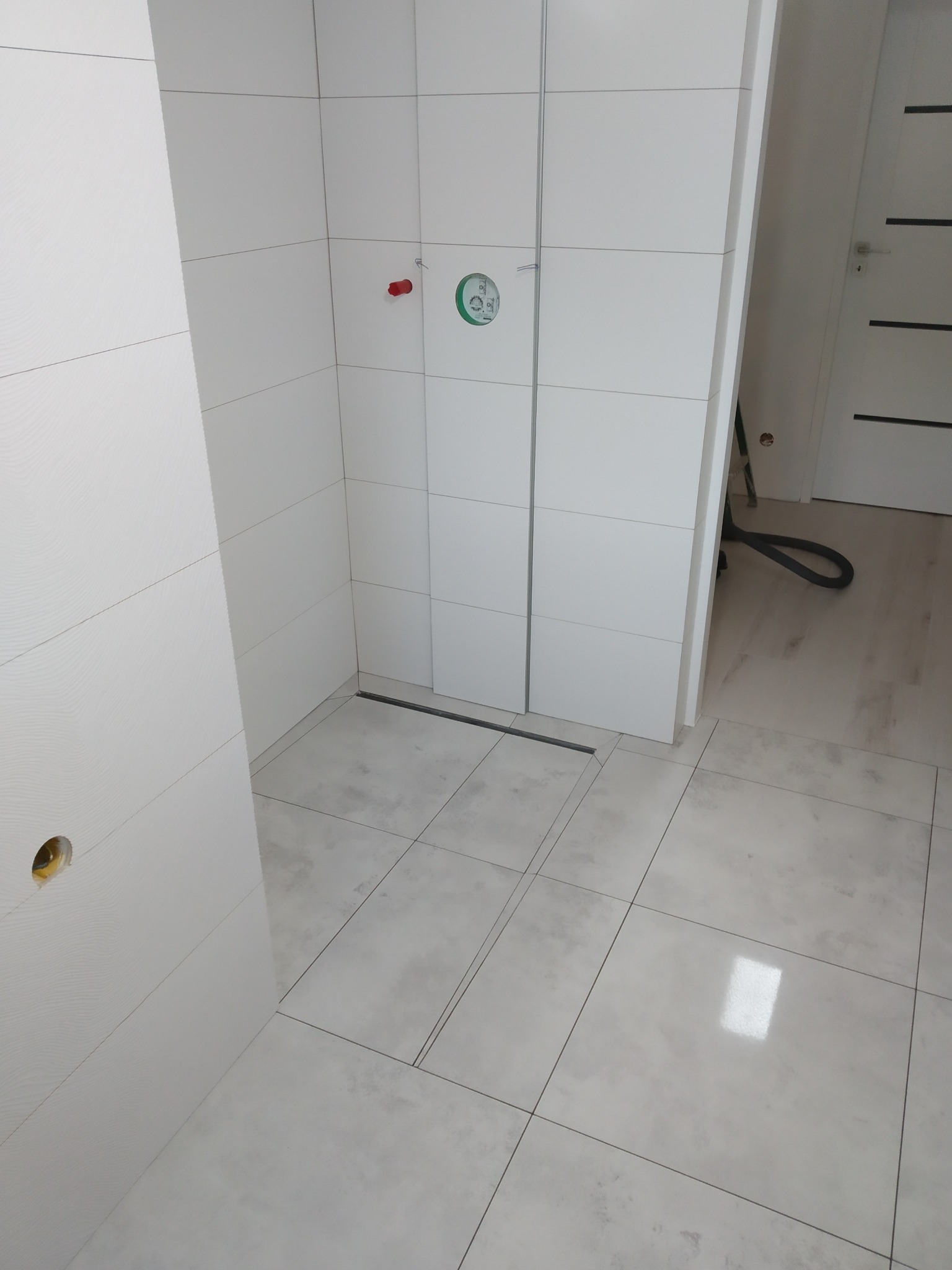 Biała, minimalistyczna łazienka z odpływem liniowym i dużymi, kwadratowymi płytkami. Widoczne przyłącza wodne i fragment odkurzacza w tle.