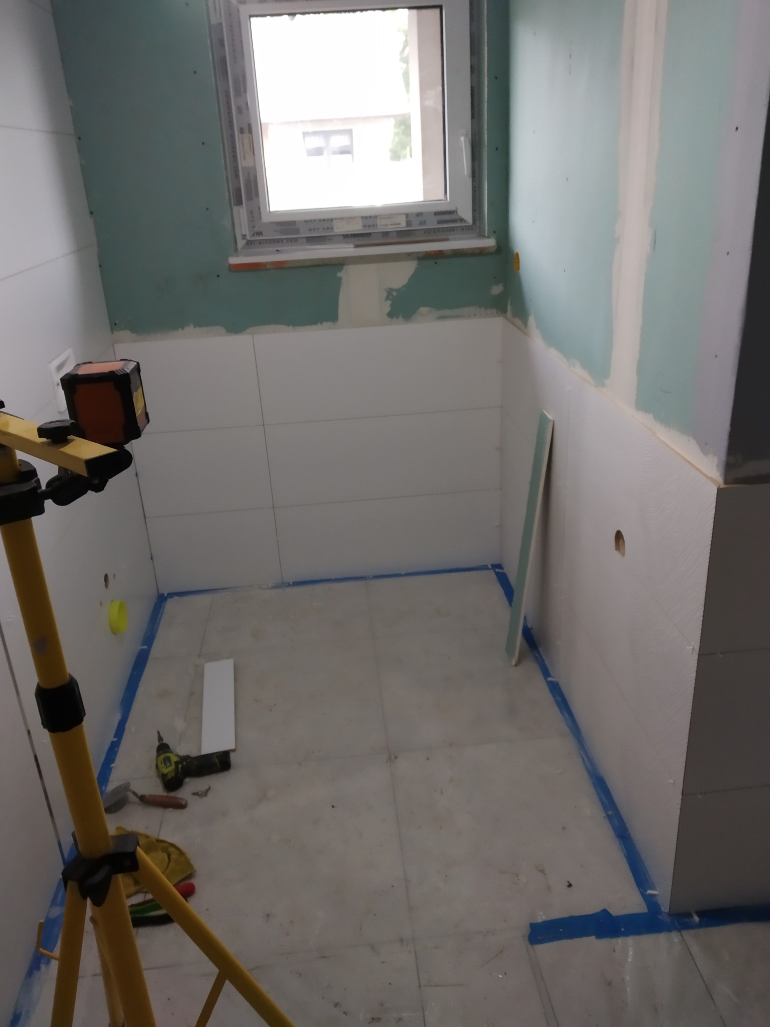 Łazienka w trakcie prac: białe płytki na ścianach do połowy wysokości, laser krzyżowy na statywie, okno w tle. Widoczne narzędzia i zabezpieczenia podłogi.