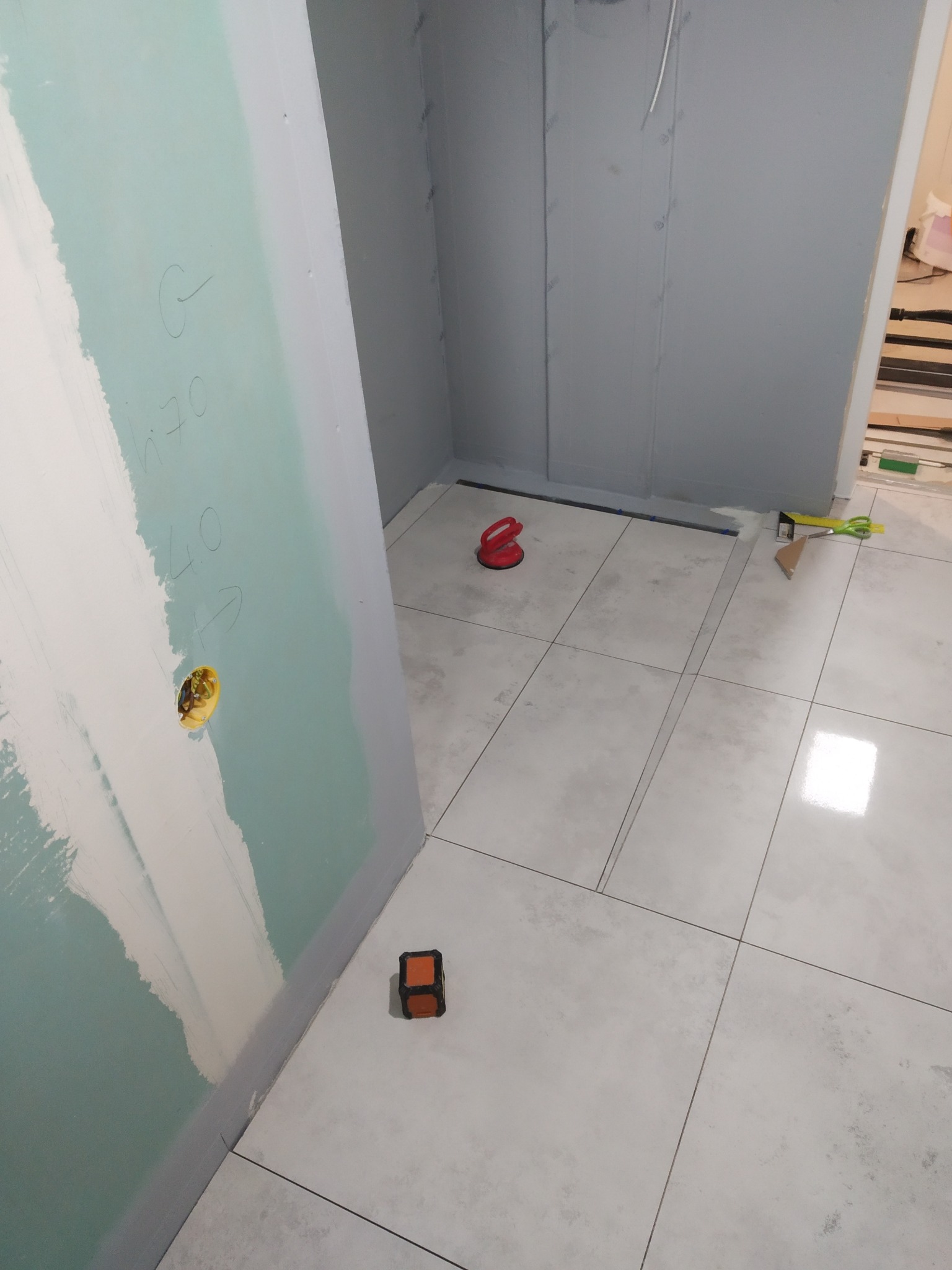 Białe płytki podłogowe w trakcie układania w rogu pomieszczenia. Na ścianach widoczne płyty gipsowo-kartonowe przygotowane do dalszych prac wykończeniowych.