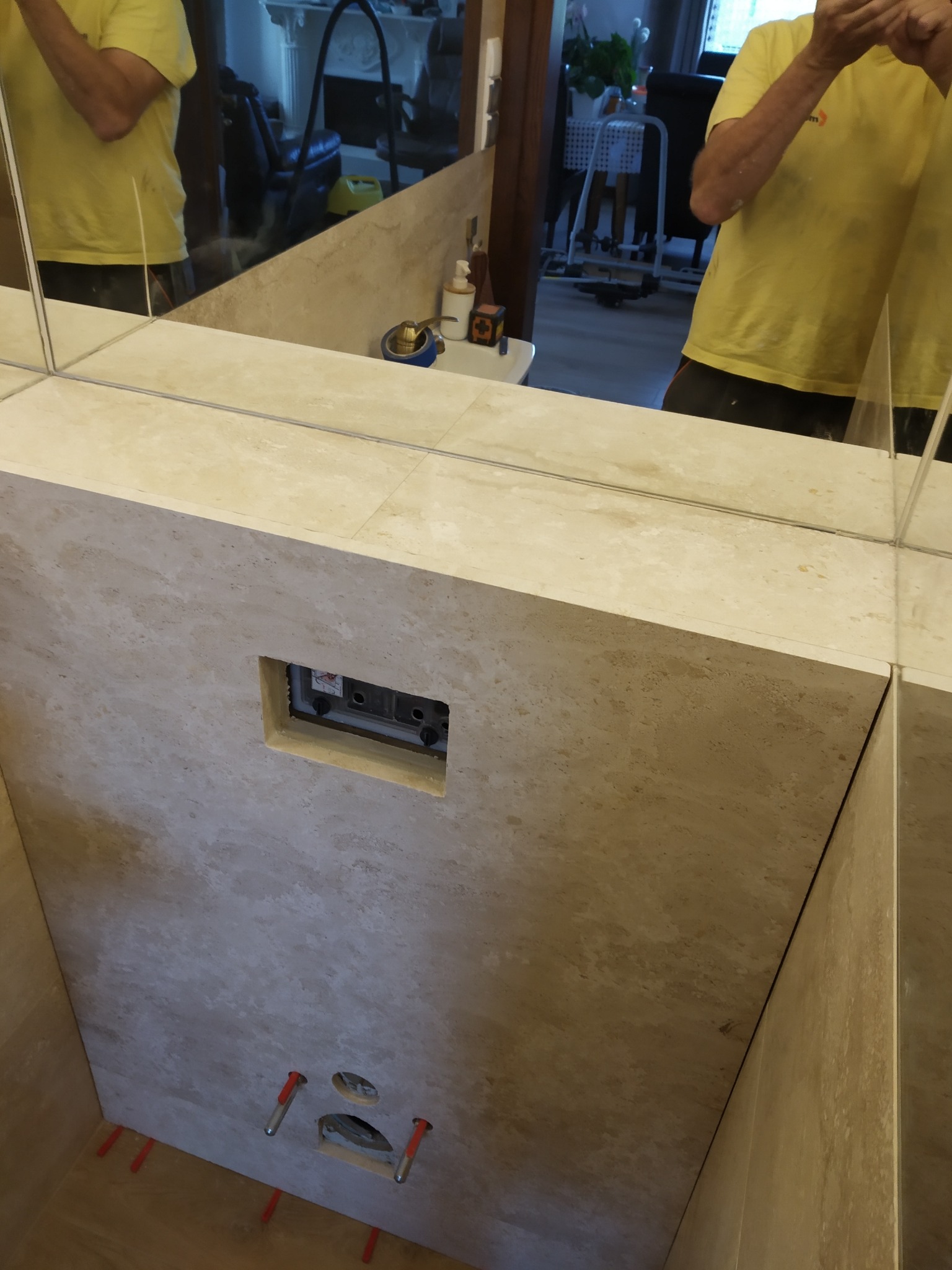 Beżowa ściana z płytek w łazience, widoczne otwory na armaturę i fragment lustra z odbiciem osoby. Czerwone kliny poziomujące płytki na podłodze.