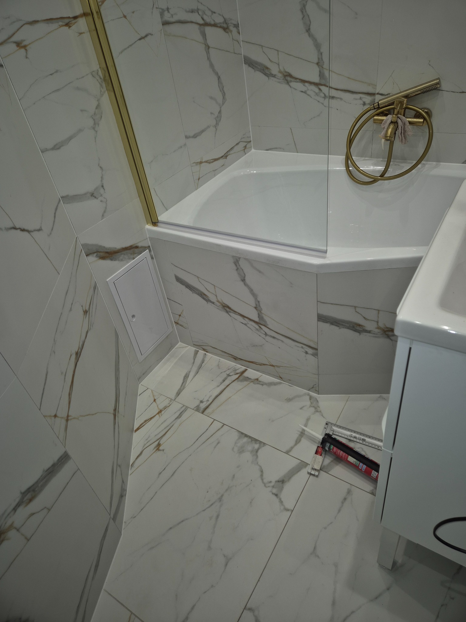 Biała łazienka z marmurowymi płytkami, złota armatura i biała wanna z parawanem. Na podłodze leży tuba z silikonem i miarka.