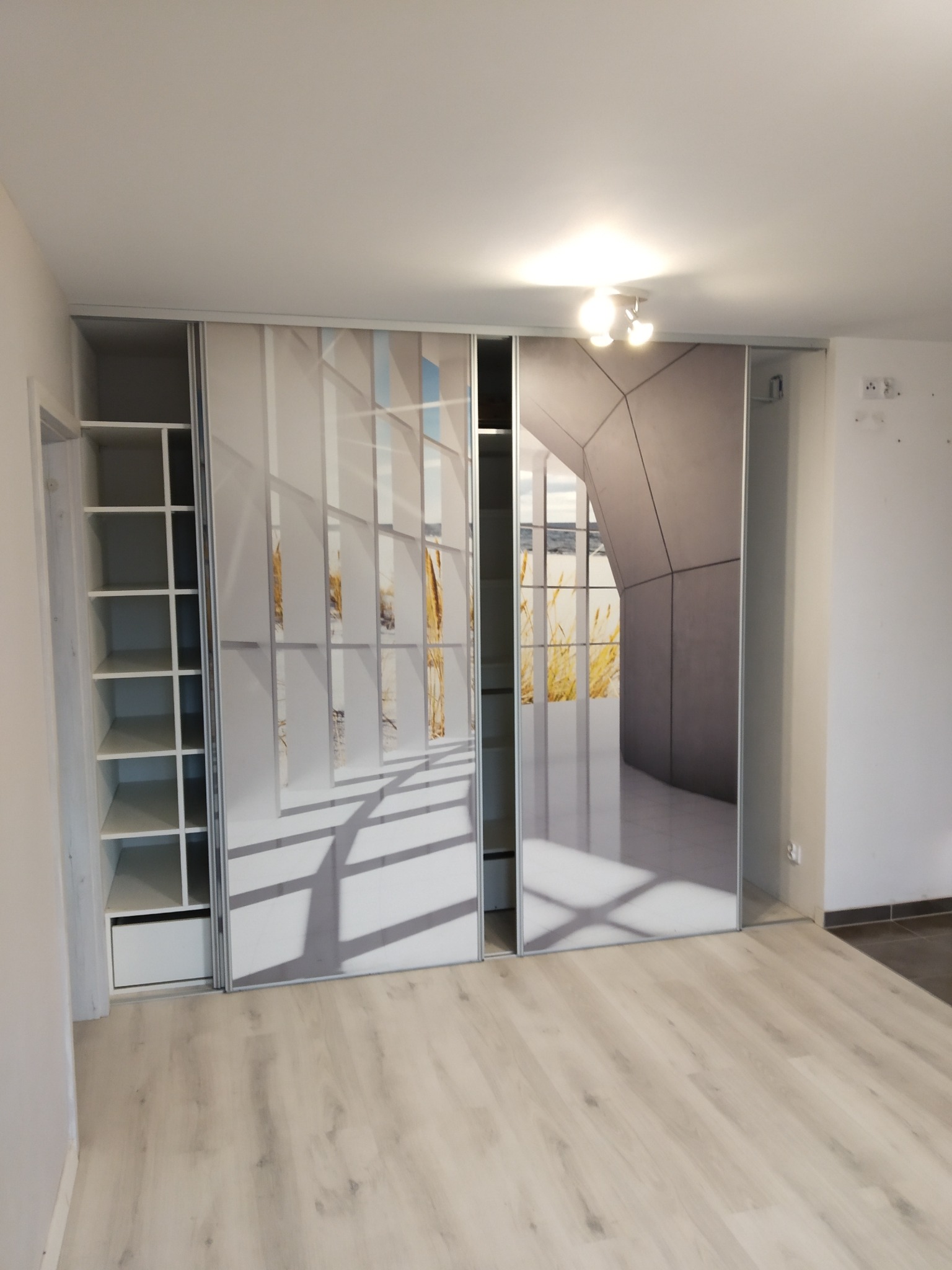 Biała szafa przesuwna z grafiką okna i krajobrazu w nowoczesnym wnętrzu. Widoczne półki po lewej stronie i panele podłogowe w jasnym odcieniu drewna.