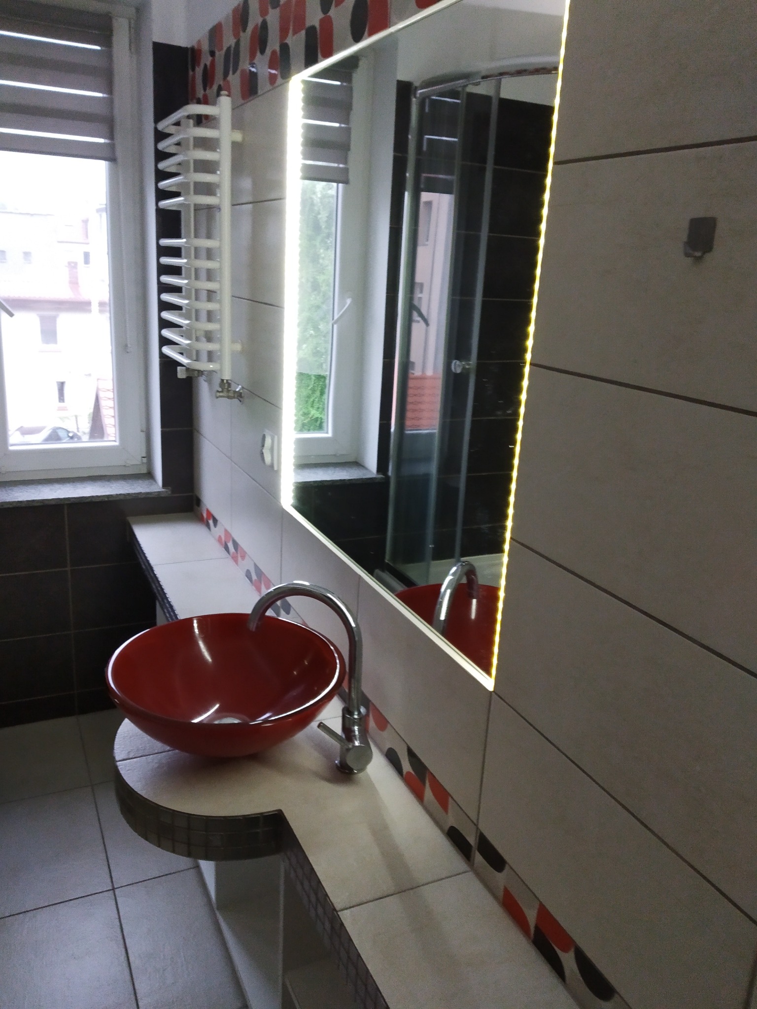 Wykończona łazienka z czerwoną umywalką na blacie z płytek, podświetlane lustro i grzejnik drabinkowy przy oknie z żaluzjami. Widoczna kabina prysznicowa.
