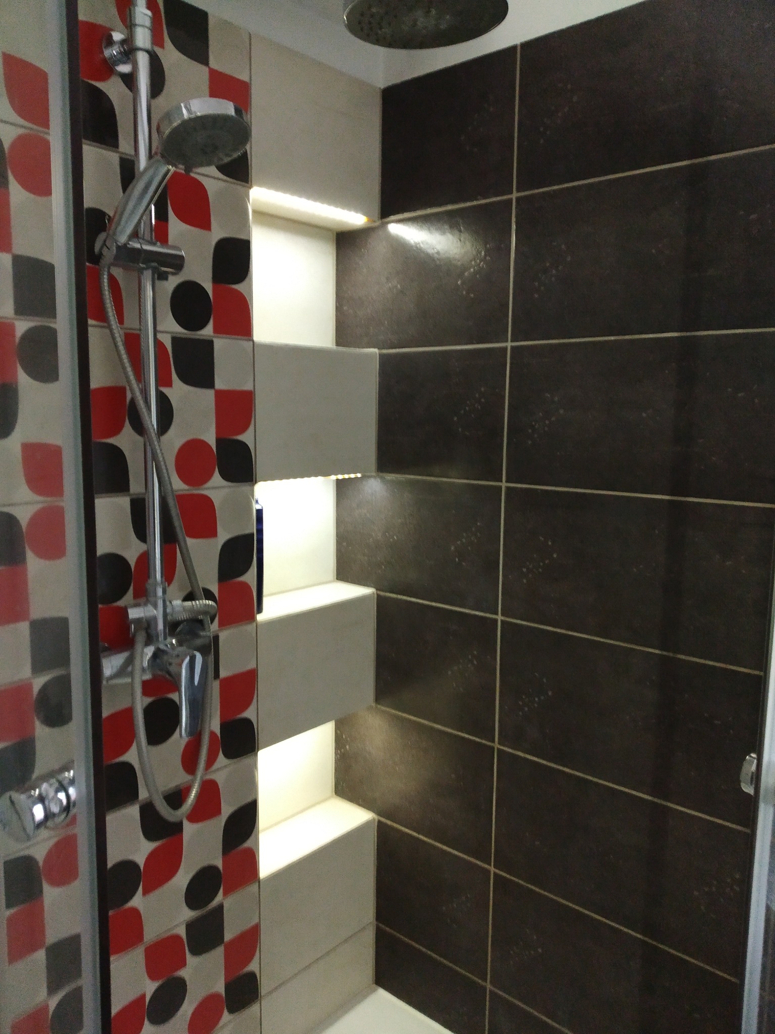 Nowoczesna kabina prysznicowa z geometrycznym wzorem płytek w odcieniach czerwieni i czerni, oraz ciemnobrązowe płytki z subtelnym oświetleniem LED.