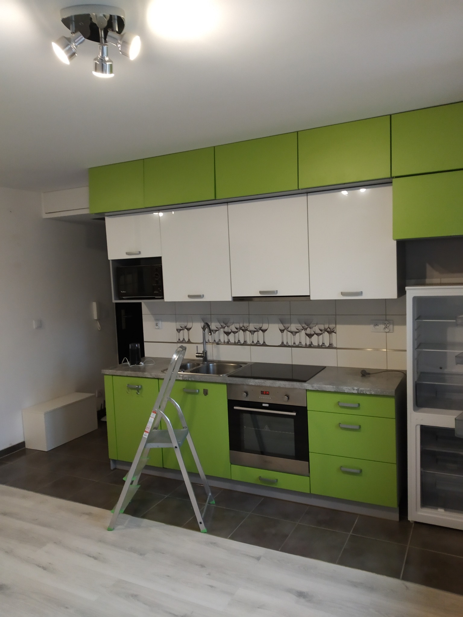 Nowoczesna kuchnia z zielonymi i białymi szafkami, srebrną drabinką i sprzętem AGD. Na ścianie płytki z motywem kieliszków. Oświetlenie sufitowe.