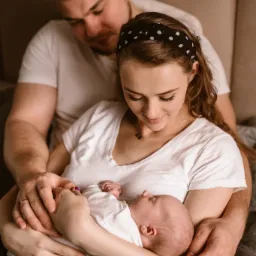 Szczęśliwa rodzina: mama z opaską w kropki trzyma śpiące niemowlę w ramionach, tata obejmuje je od tyłu, wszyscy ubrani w białe koszulki, ciepłe, naturalne światło.