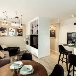 Nowoczesne wnętrze mieszkania z otwartą kuchnią, brązową sofą, okrągłym drewnianym stołem z zastawą i czarnymi hokerami przy wyspie.