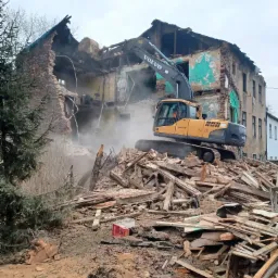 Żółta koparka Volvo na stercie gruzu podczas rozbiórki częściowo zburzonego, dwupiętrowego budynku z cegły i tynku w pochmurny dzień.