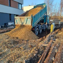 Ciężarówka z podniesioną skrzynią wyładowuje żwir na przygotowany teren budowy, widoczne pręty zbrojeniowe i wytyczone granice fundamentów.