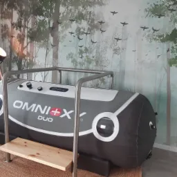 Komora hiperbaryczna Omni+X Duo z metalową ramą i drewnianym stopniem, ustawiona na okrągłym dywanie, z lampą na trójnogu i tapetą z motywem leśnym w tle.