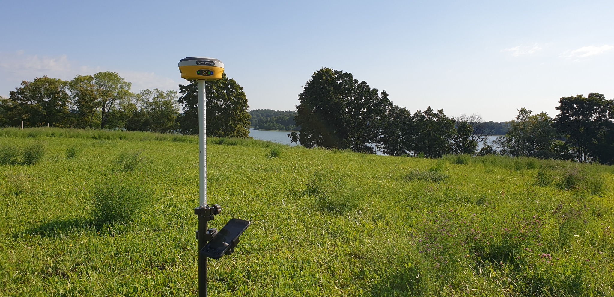 Żółty odbiornik GNSS na tyczce w trawie, w tle jezioro i drzewa. Urządzenie pomiarowe ART-GEO na tle zieleni i błękitu. Sprzęt do pomiarów geodezyjnych w terenie.
