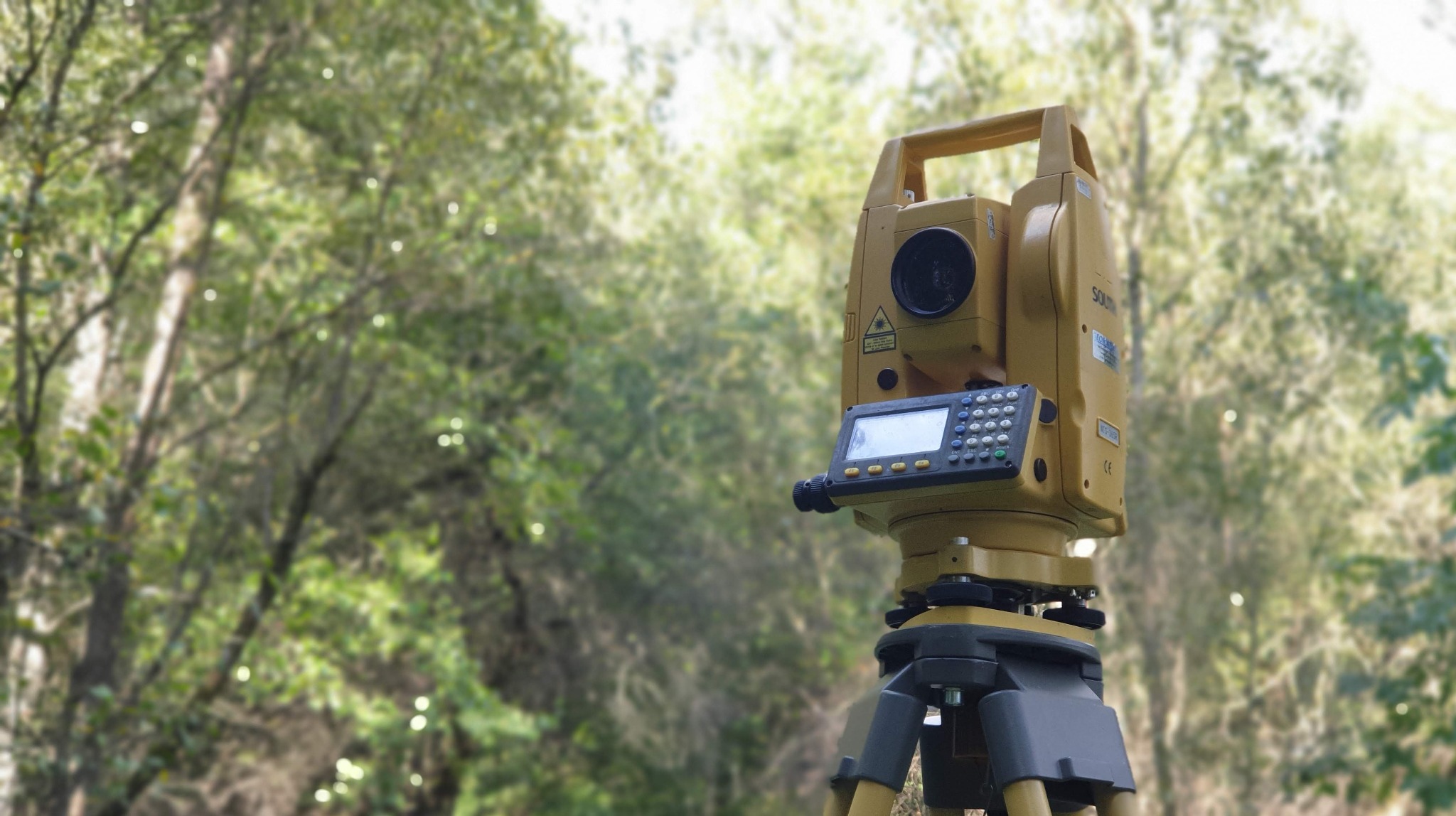 Żółty teodolit na statywie w terenie zielonym, ustawiony do pomiarów. Widoczne drzewa w tle, sprzęt gotowy do pracy.