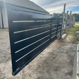 Nowoczesna brama wjazdowa przesuwna, antracytowa, z poziomymi szczelinami, na tle domu z szarą dachówką i częściowo widocznym ogrodzeniem z drewnianych słupków.