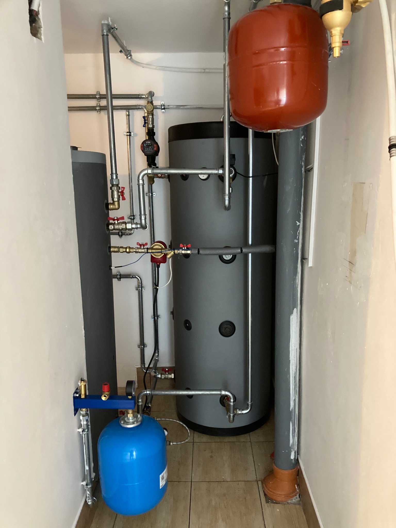 Dwa szare zbiorniki akumulacyjne ciepła o różnej wielkości, połączone rurami z zaworami, z czerwonym i niebieskim zbiornikiem wyrównawczym, zainstalowane w pomieszczeniu technicznym z beżowymi...