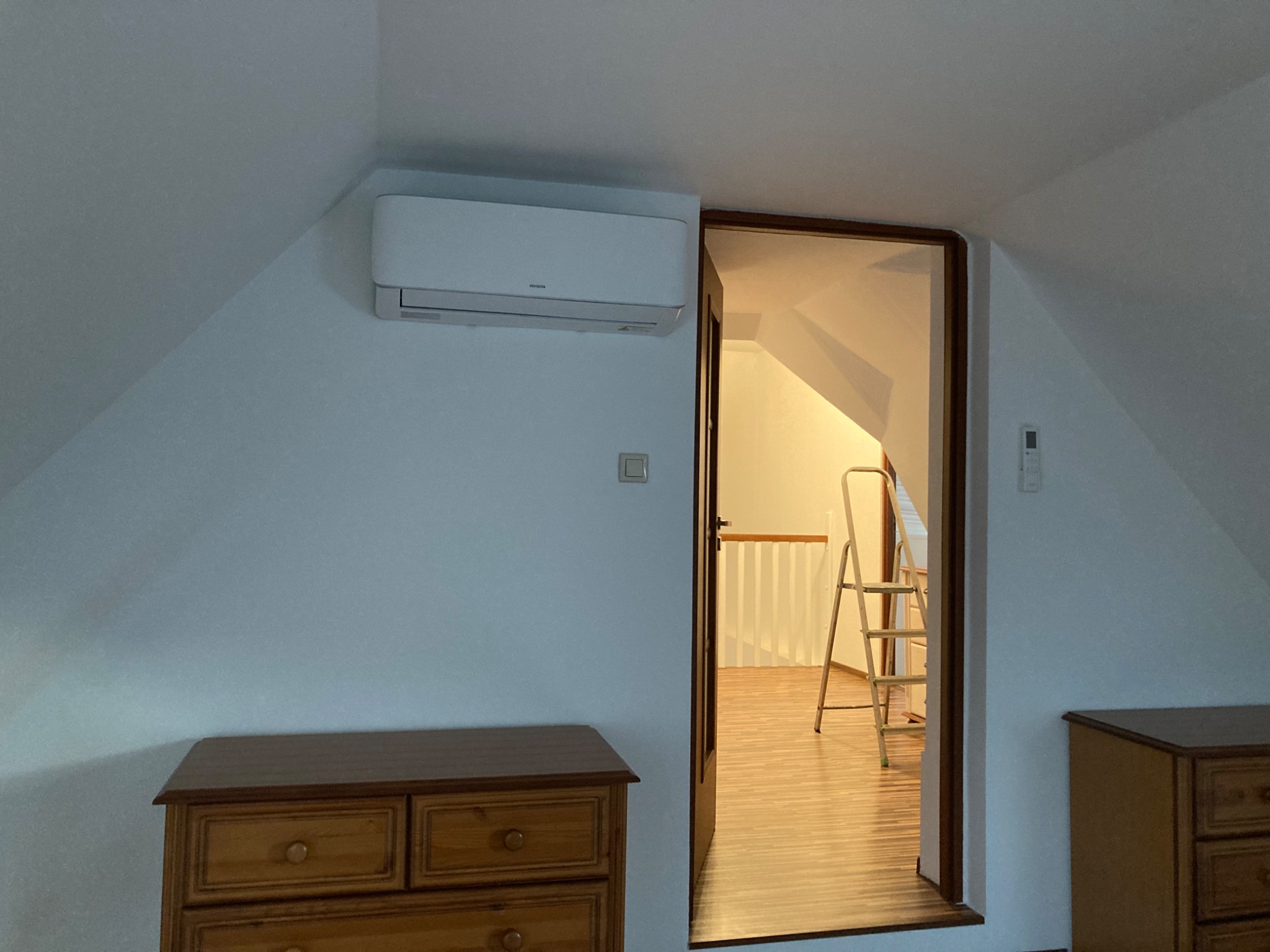 Biały klimatyzator zamontowany na skośnej ścianie poddasza, widoczny obok otwartych drzwi z drabinką w tle.
