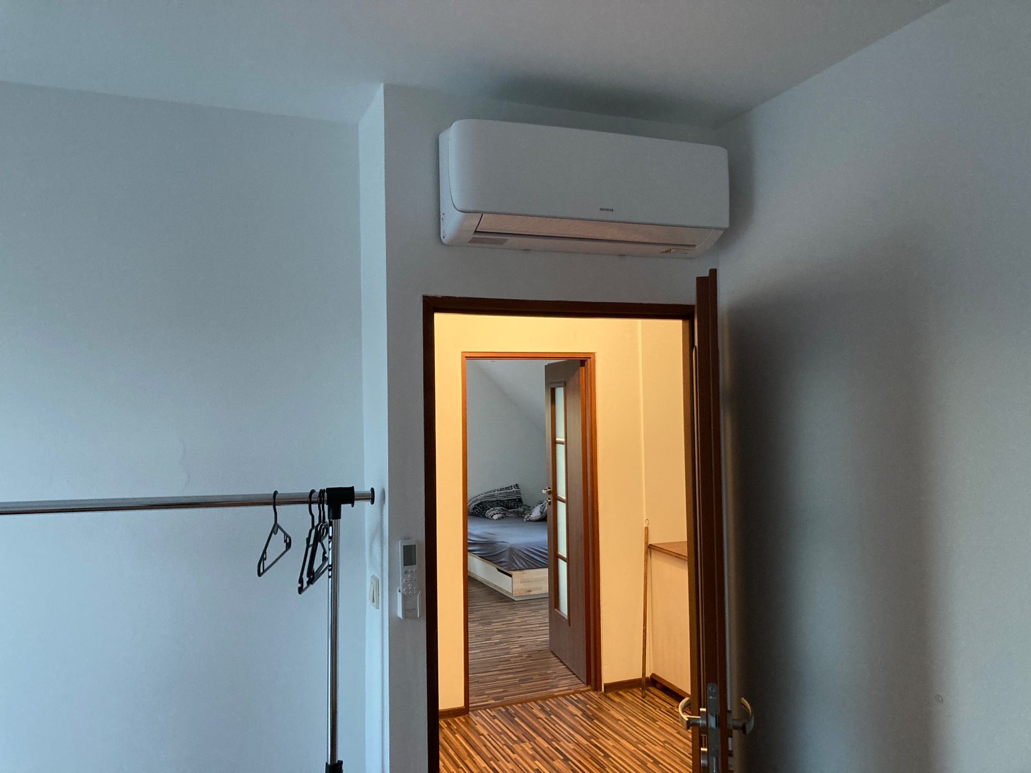 Biały klimatyzator ścienny zamontowany nad otwartymi drzwiami do sypialni z widocznym łóżkiem, na ścianie wieszak na ubrania.