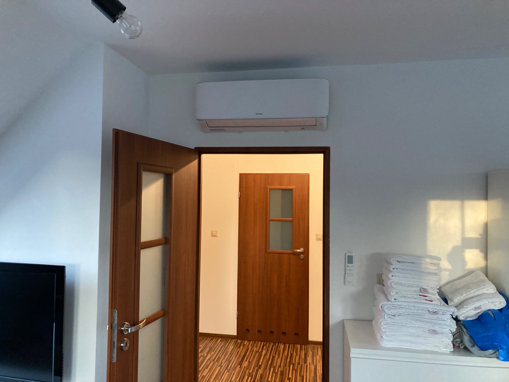 Biała jednostka klimatyzacyjna zamontowana nad otwartymi, drewnianymi drzwiami w pokoju z stertą białych ręczników i nieosłoniętą żarówką.