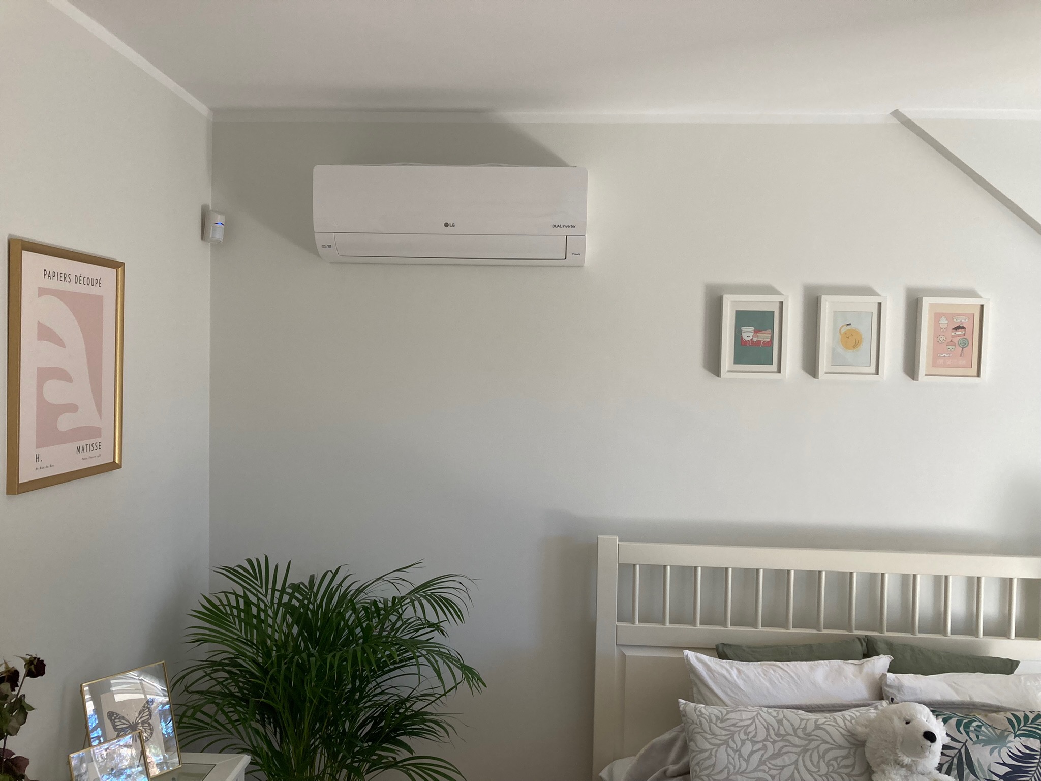 Biała jednostka wewnętrzna klimatyzatora LG zamontowana na ścianie w sypialni, widoczne trzy małe obrazki w białych ramkach oraz plakat Matisse, roślina doniczkowa i zagłówek łóżka.
