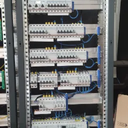 Rozdzielnia elektryczna z licznymi zabezpieczeniami nadprądowymi, modułami różnicowoprądowymi i okablowaniem w kolorach niebieskim i żółto-zielonym, zamontowana w metalowej szafie rack.