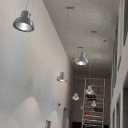 Korytarz biurowy z zamontowanymi srebrnymi lampami wiszącymi, drabina rusztowaniowa w tle, widoczne puszki elektryczne na ścianie.