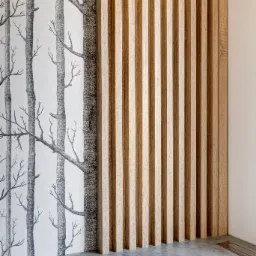 Drewniane lamele pionowe tworzące dekoracyjną ściankę działową, na tle tapeta z motywem drzew, szara podłoga.