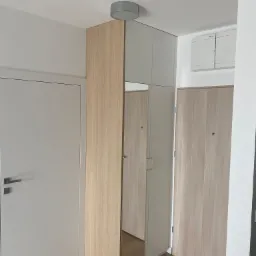 Wysoka szafa z lustrem wkomponowana we wnękę, z frontem w kolorze jasnego drewna, sąsiadująca z białymi drzwiami w minimalistycznym wnętrzu.