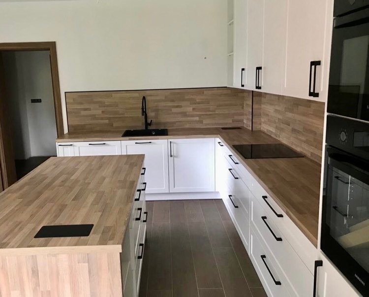 Nowoczesna kuchnia z białymi szafkami, drewnianymi blatami i czarnymi uchwytami, widoczna wyspa kuchenna z drewnianym blatem i wbudowanym gniazdkiem.