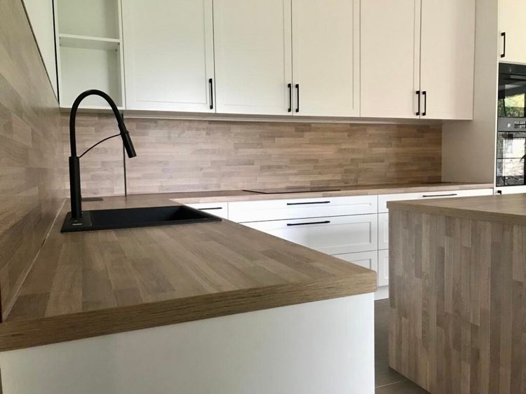 Nowoczesna kuchnia z białymi szafkami i drewnianym blatem oraz wyspą, czarna bateria kuchenna, drewniana mozaika na ścianie.