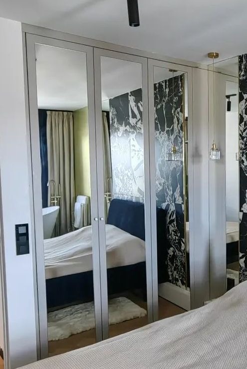 Czterodrzwiowa szafa z lustrzanymi frontami w sypialni, odbijająca łazienkę z wanną i marmurowe akcenty dekoracyjne.
