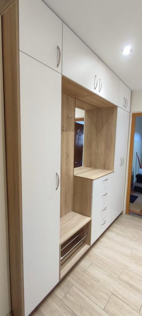 Biało-drewniana zabudowa przedpokoju z szafkami, szufladami, siedziskiem i lustrem, widok na korytarz z drzwiami.